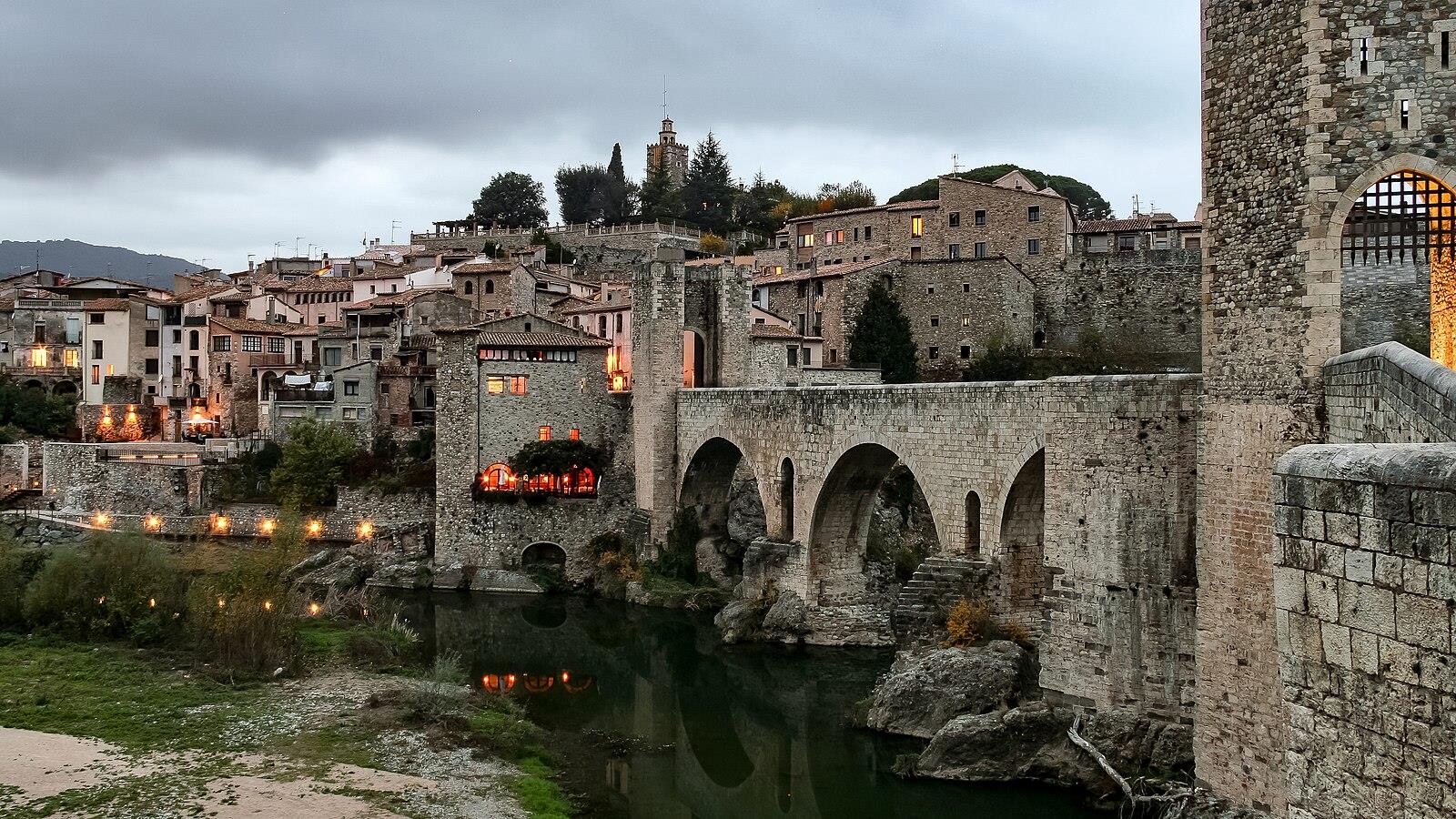 Besalú al atardecer con las luces del puente reflejadas en el río Fluvià y el casco histórico medieval al fondo
