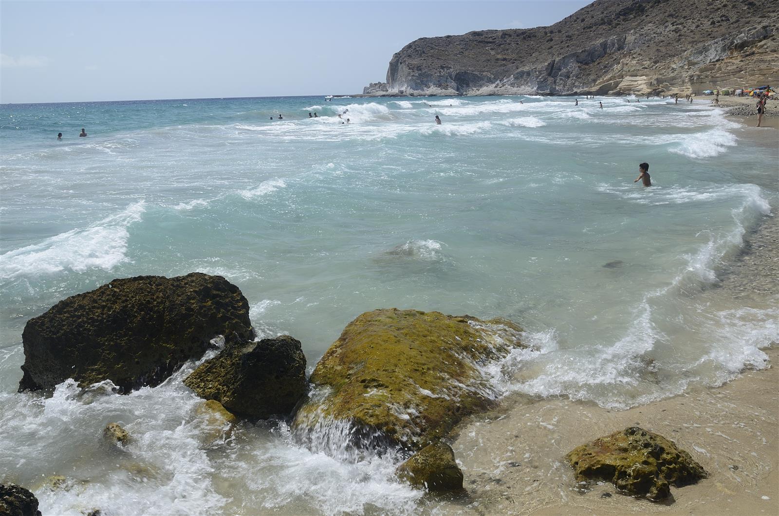 Playa de arena oscura y oleaje turquesa en Cala del Plomo con bañistas y acantilados al fondo