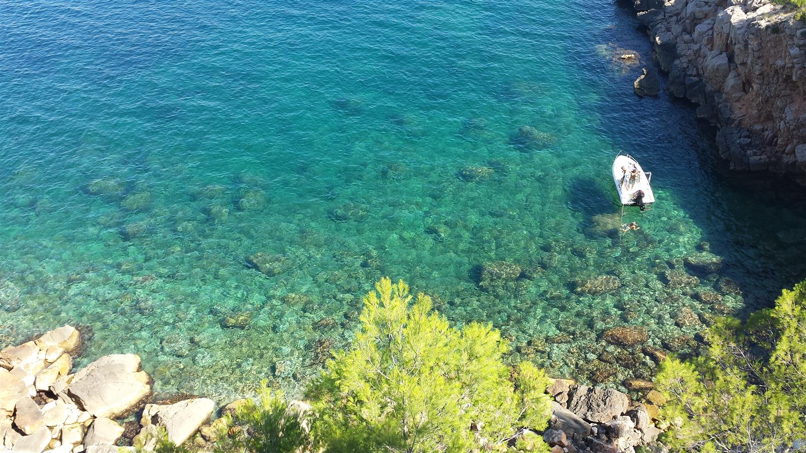 Agua turquesa cristalina de Cala Montgó con roca mediterránea, pinos en primer plano y un barco anclado, Costa Brava, Girona