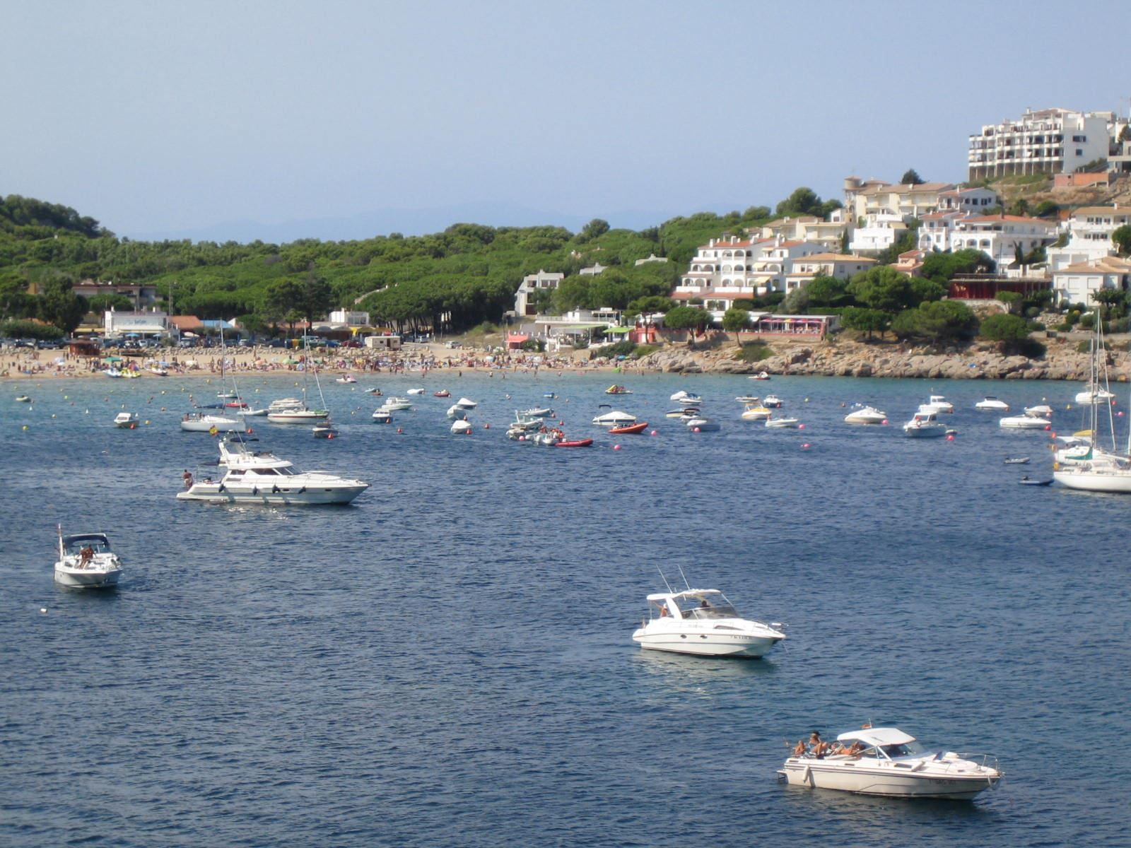 Playa de Cala Montgó en verano vista desde el mar con bañistas, barcos, pinos y la urbanización de L'Escala al fondo