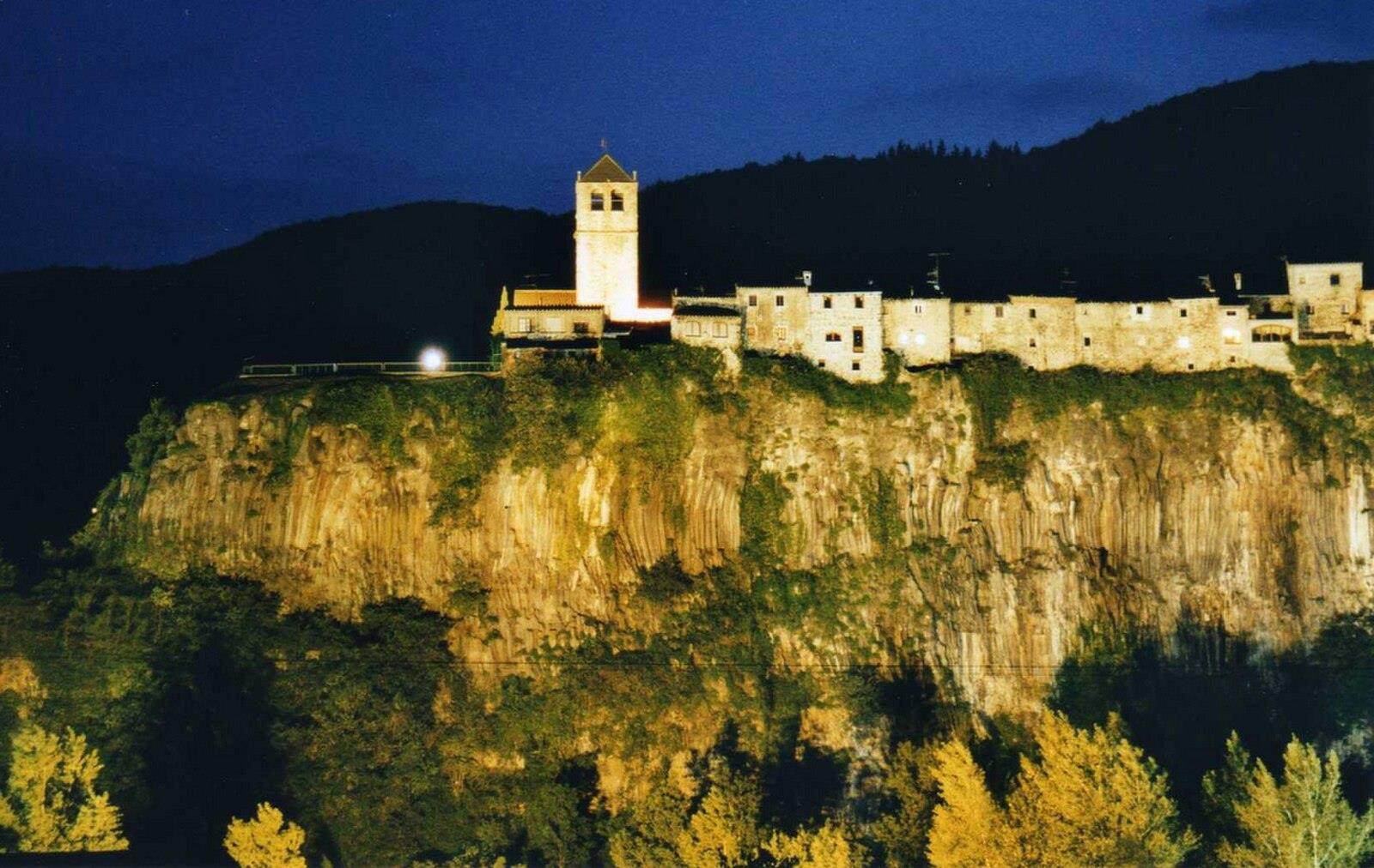 Castellfollit de la Roca iluminado de noche sobre el risco basáltico, con la luz artificial dorada reflejando en las columnas de basalto