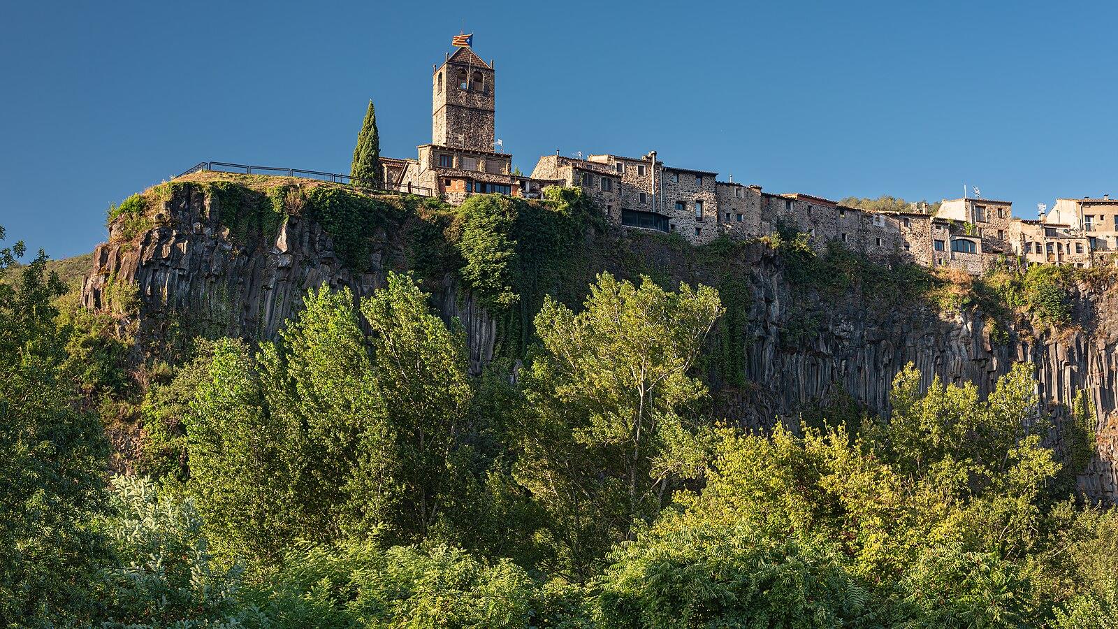 Castellfollit de la Roca visto desde el exterior: el pueblo encaramado sobre el risco basáltico volcánico con la torre de la iglesia sobre el acantilado, cielo azul y vegetación verde en la ribera del Fluvià