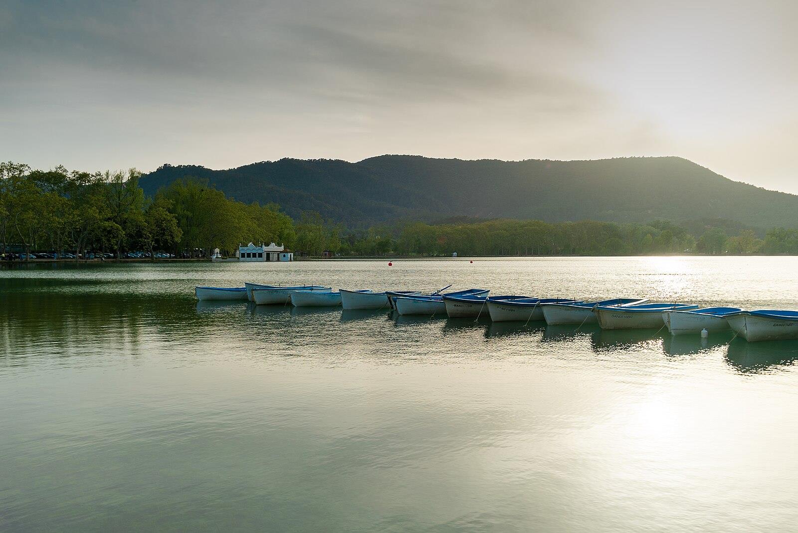 Barcas de remo blancas y azules amarradas en el Estany de Banyoles al amanecer, con el lago y las montañas de fondo