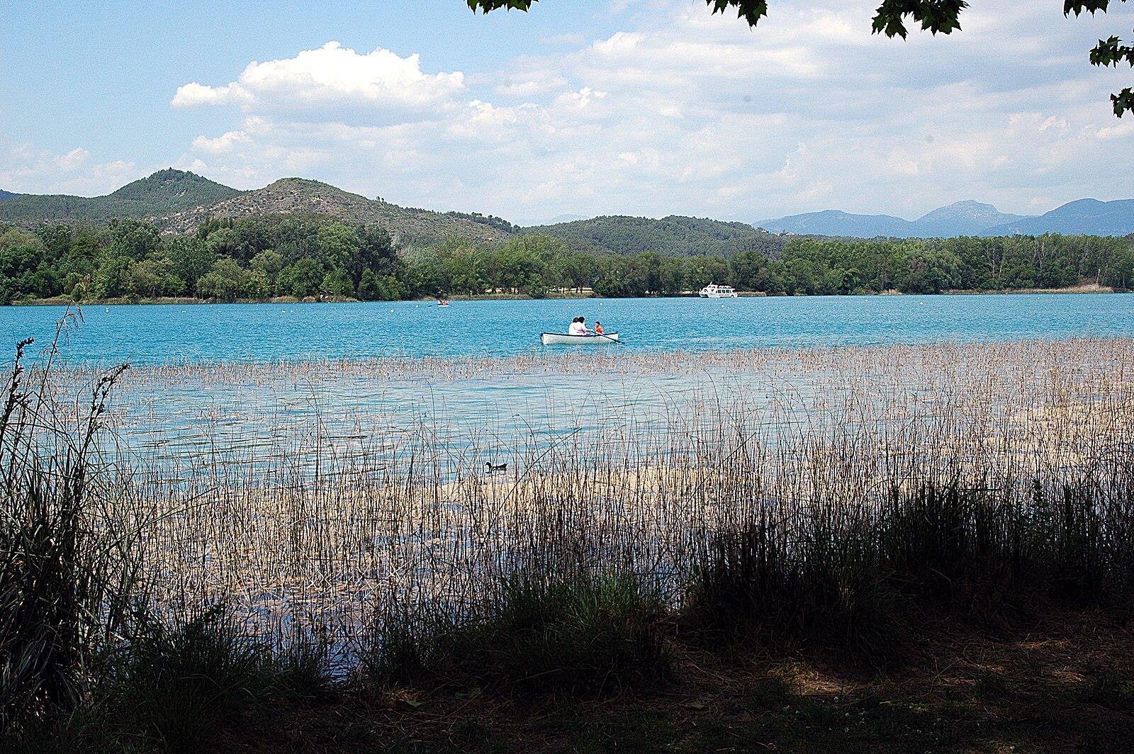 Vista del Estany de Banyoles desde los juncos con una canoa en el agua turquesa y las montañas de la Garrotxa al fondo