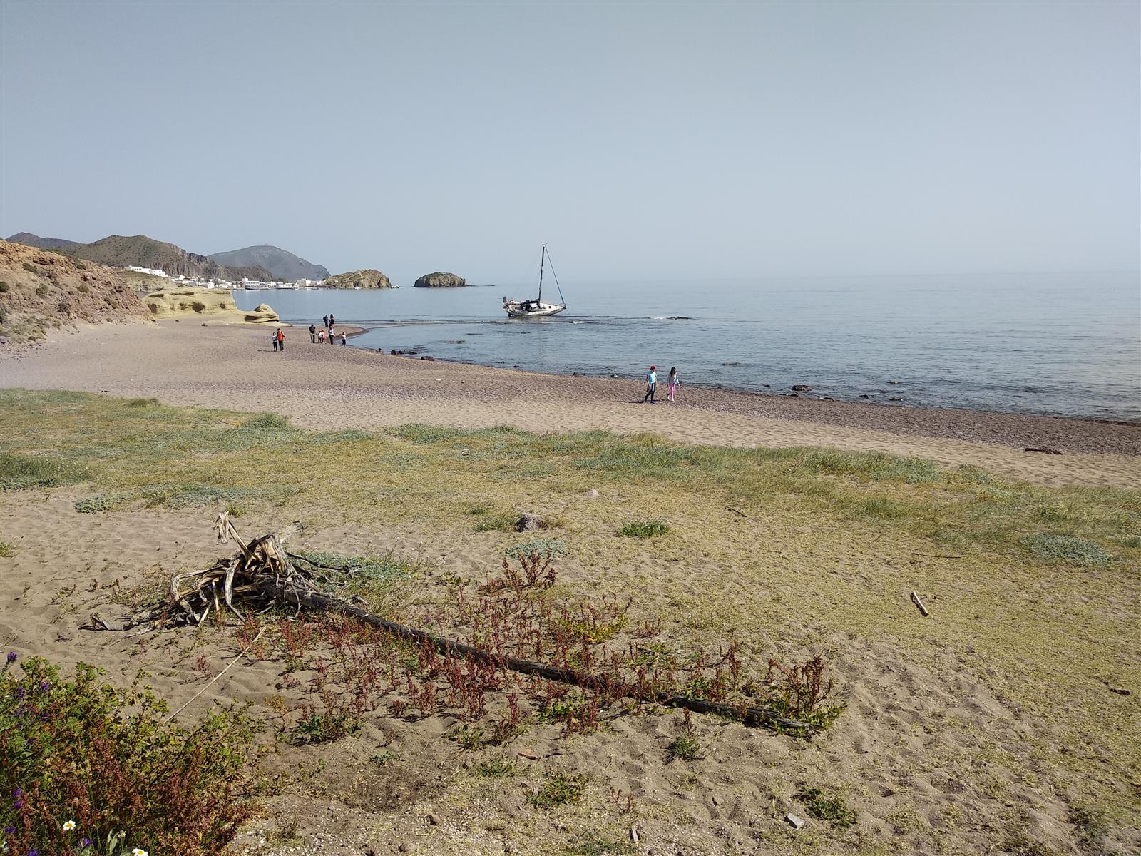 Playa del Arco en Los Escullos a nivel del suelo, con personas paseando, un velero encallado cerca de la orilla y La Isleta del Moro al fondo
