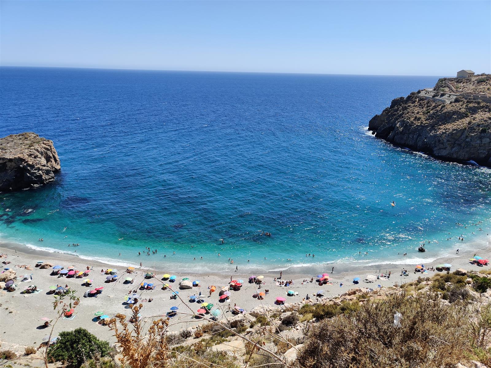 Vista cenital de la Playa de La Rijana desde el acantilado, con bañistas en el agua y sombrillas de colores en la orilla