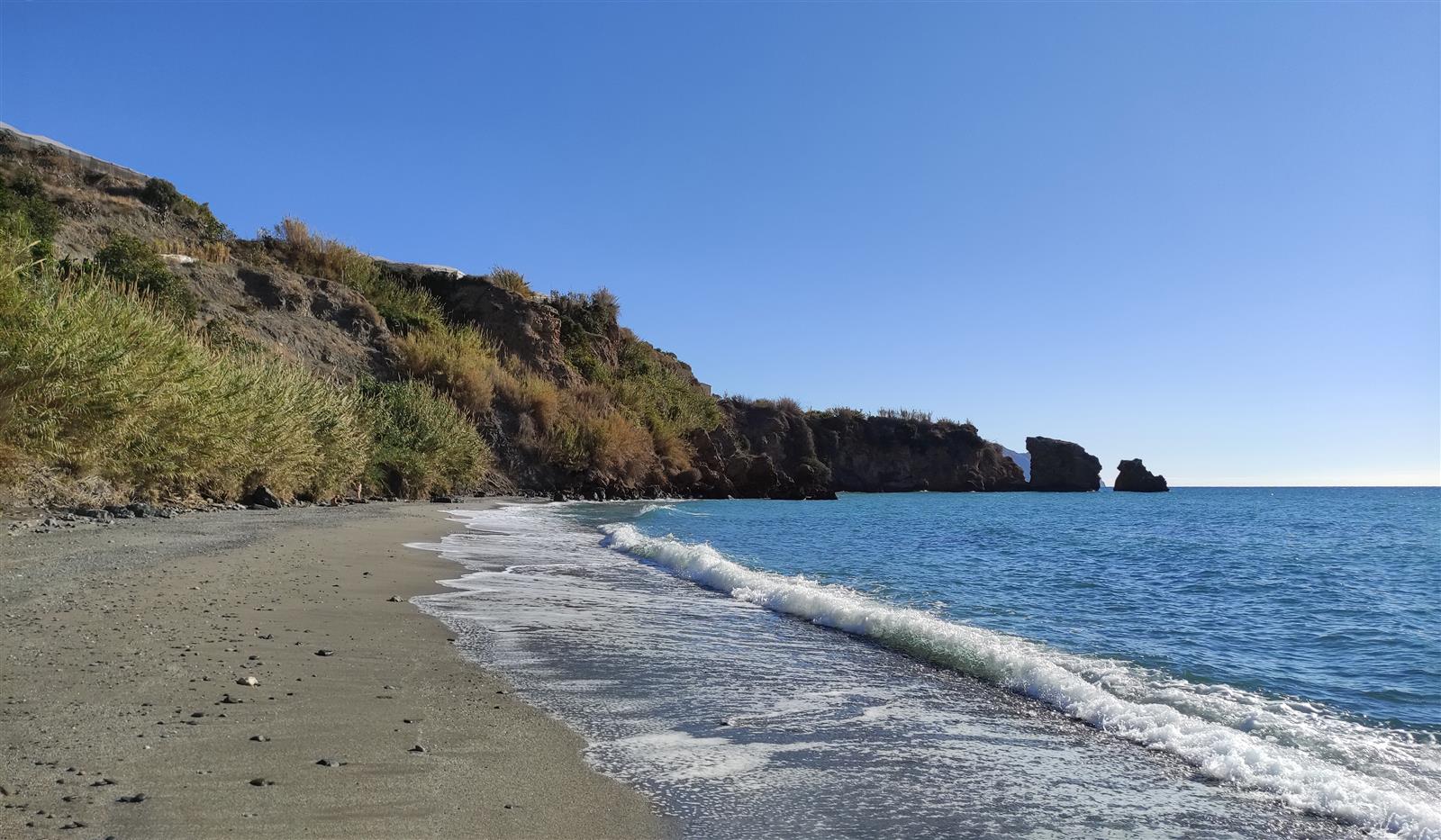Caleta de Maro con agua turquesa, oleaje suave y acantilados al fondo en un día de cielo azul claro