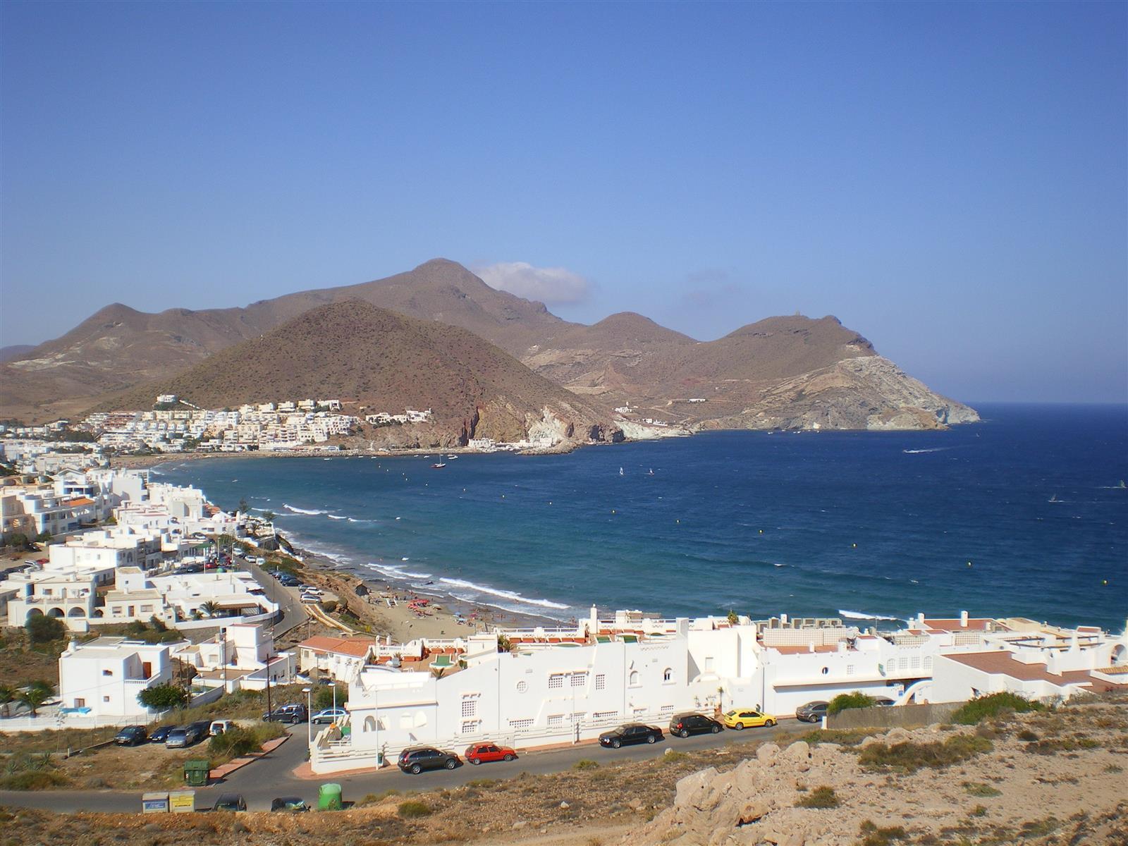 Vista aérea de la bahía de San José con el pueblo blanco, la playa curva y la sierra volcánica de Cabo de Gata al fondo, Almería