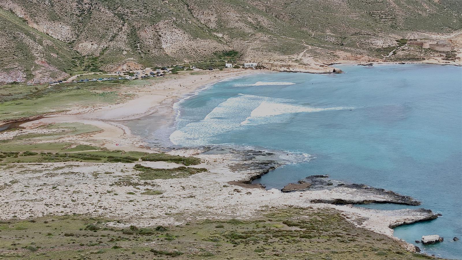 Vista aérea de El Playazo de Rodalquilar mostrando la bahía completa, el aparcamiento, el Castillo de San Ramón en lo alto y la roca volcánica de la costa