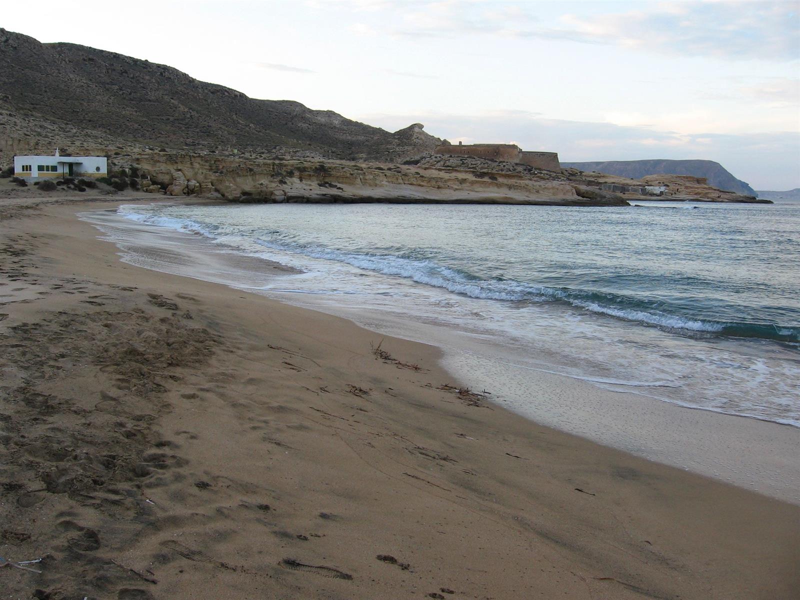 Atardecer en El Playazo de Rodalquilar con la arena, el mar con pequeñas olas y el Castillo de San Ramón visible sobre el acantilado al fondo