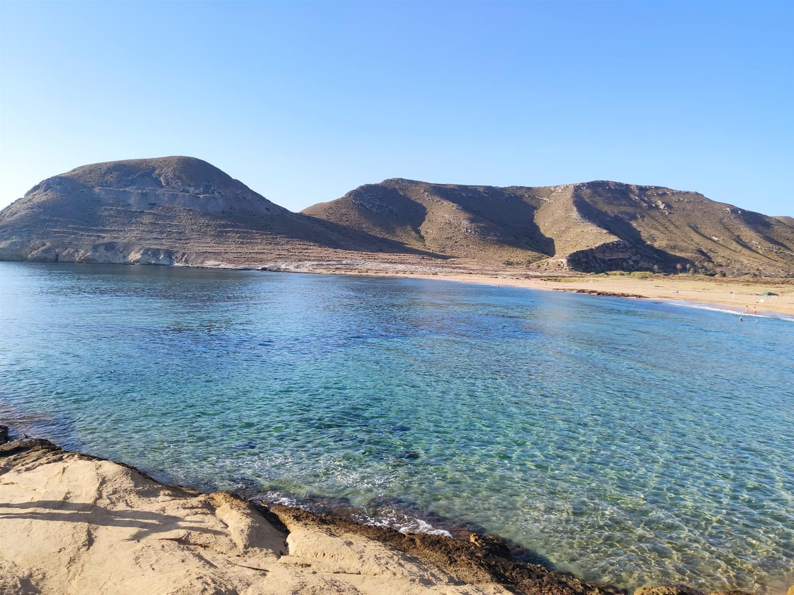 Agua turquesa cristalina en primer plano en El Playazo de Rodalquilar, con la playa de arena y el perfil del Cerro de la Molata al fondo bajo cielo azul