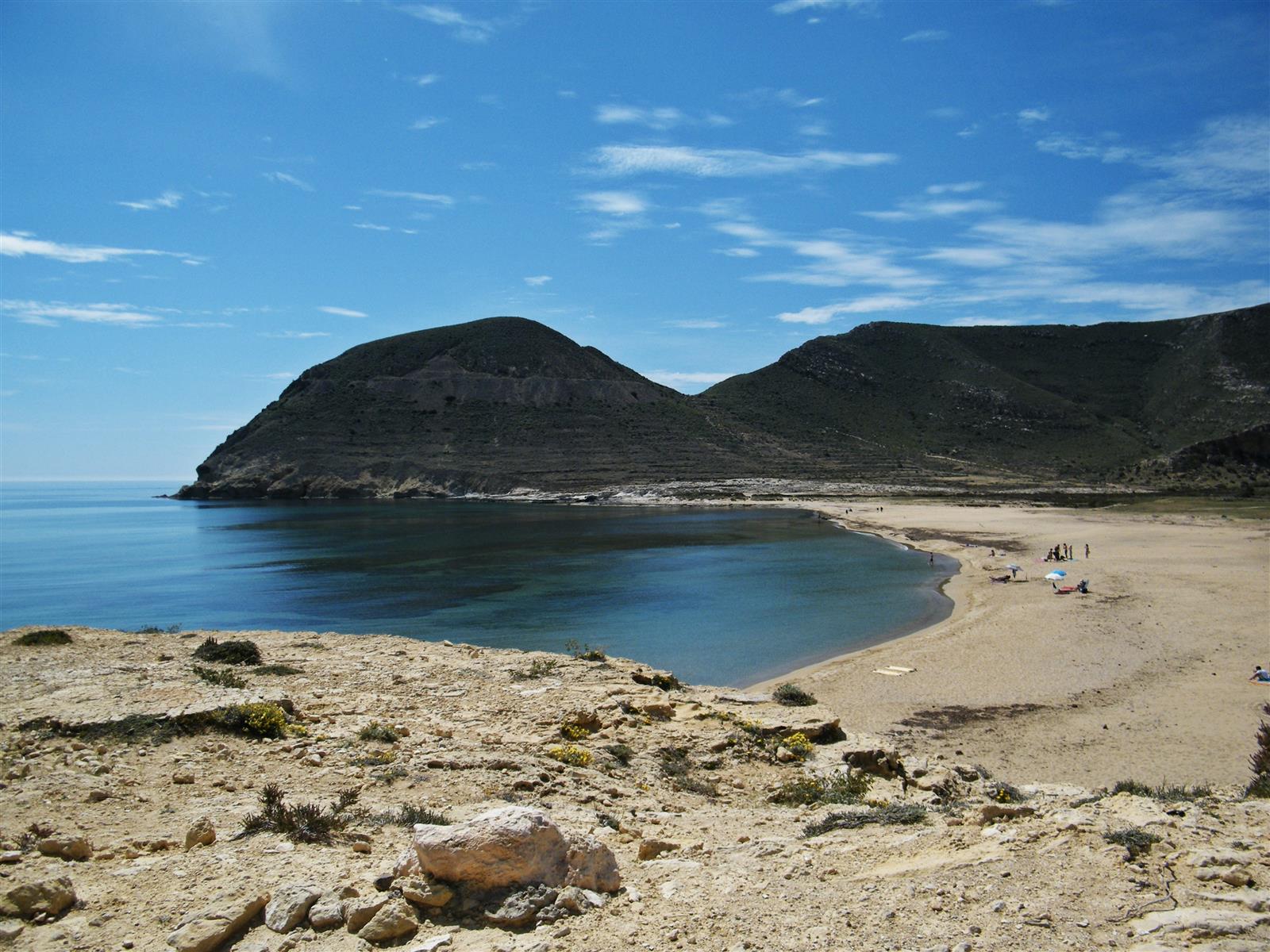 Vista panorámica elevada de El Playazo de Rodalquilar con la bahía en arco, arena dorada, mar azul y el Cerro de la Molata al fondo bajo cielo azul