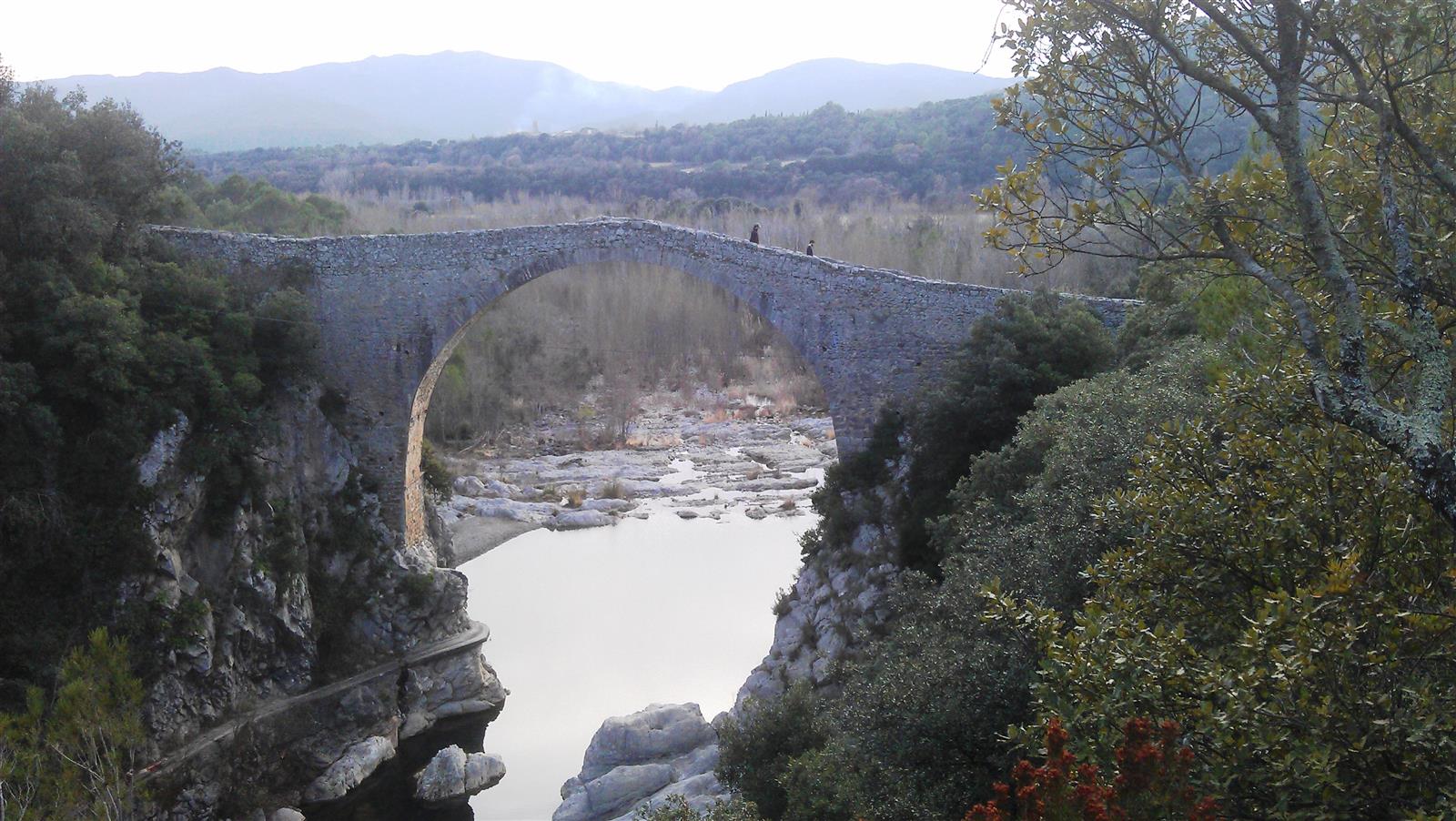Vista lateral del Pont de Llierca desde una posición elevada: el puente de piedra completo con el cañón del río Llierca y el paisaje de La Garrotxa al fondo