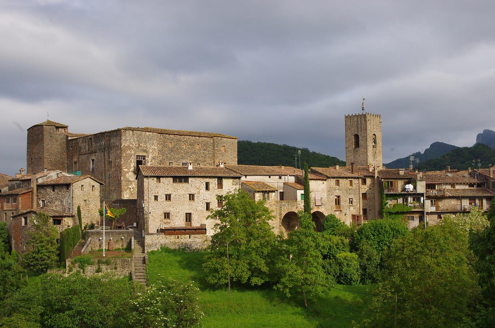 Vista general de Santa Pau: castillo a la izquierda, torre campanario a la derecha y casco histórico de piedra con los volcanes de La Garrotxa al fondo