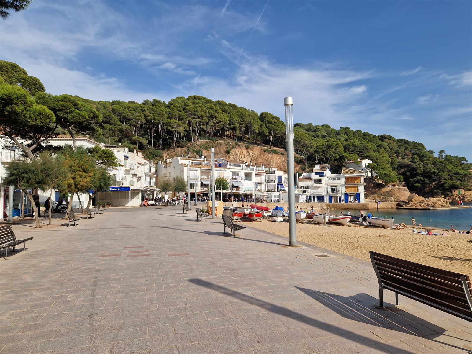 Paseo marítimo de Tamariu con bancos, playa a la derecha y pueblo de casas blancas entre pinos, Girona