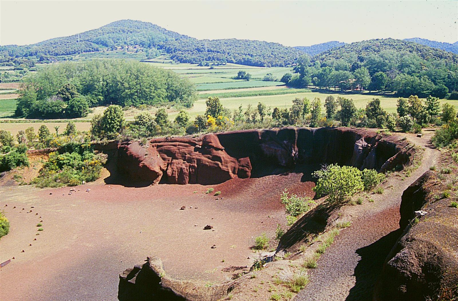 Vista desde lo alto del Volcán Croscat: tierra volcánica rojiza en primer plano y paisaje agrícola de La Garrotxa con otras colinas volcánicas al fondo