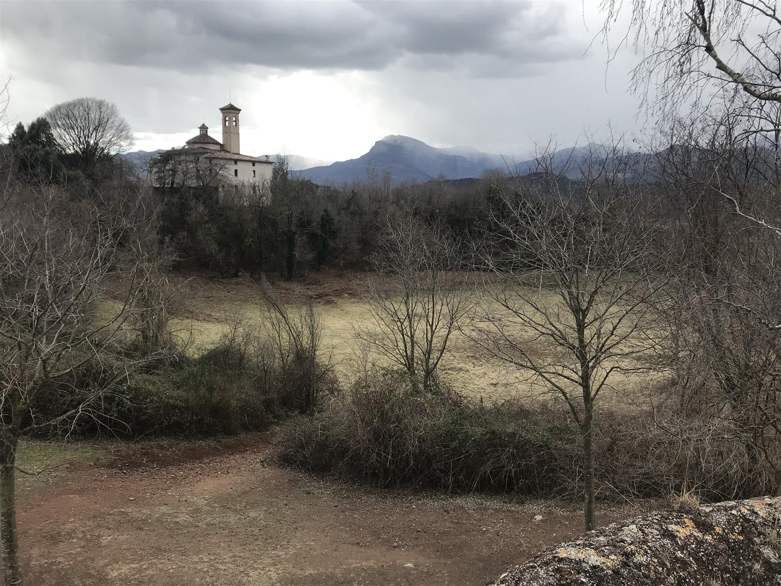 Vista interior del cráter del Volcán Montsacopa en 2024: suelo volcánico rojizo, sendero y la iglesia de Sant Francesc con montañas al fondo bajo cielo nublado